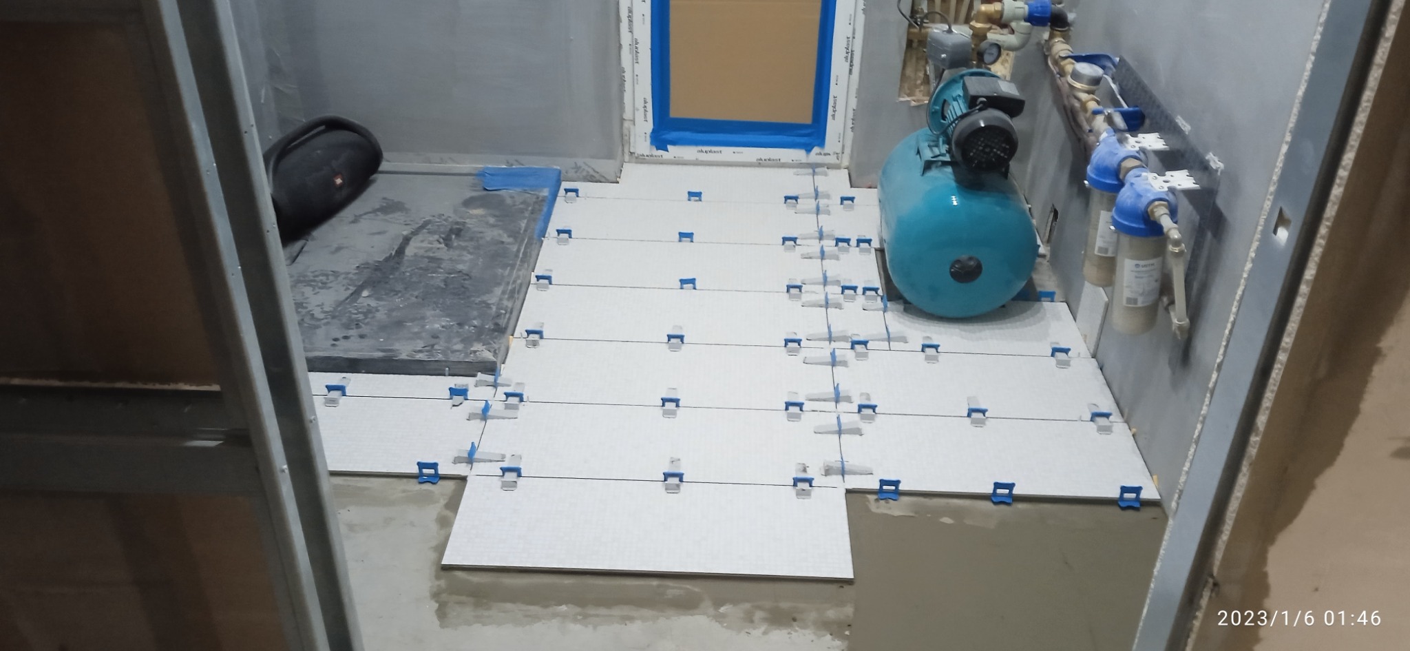 Układanie jasnych płytek podłogowych w łazience z użyciem klipsów poziomujących, widoczny zbiornik hydroforu i filtry wody na ścianie.