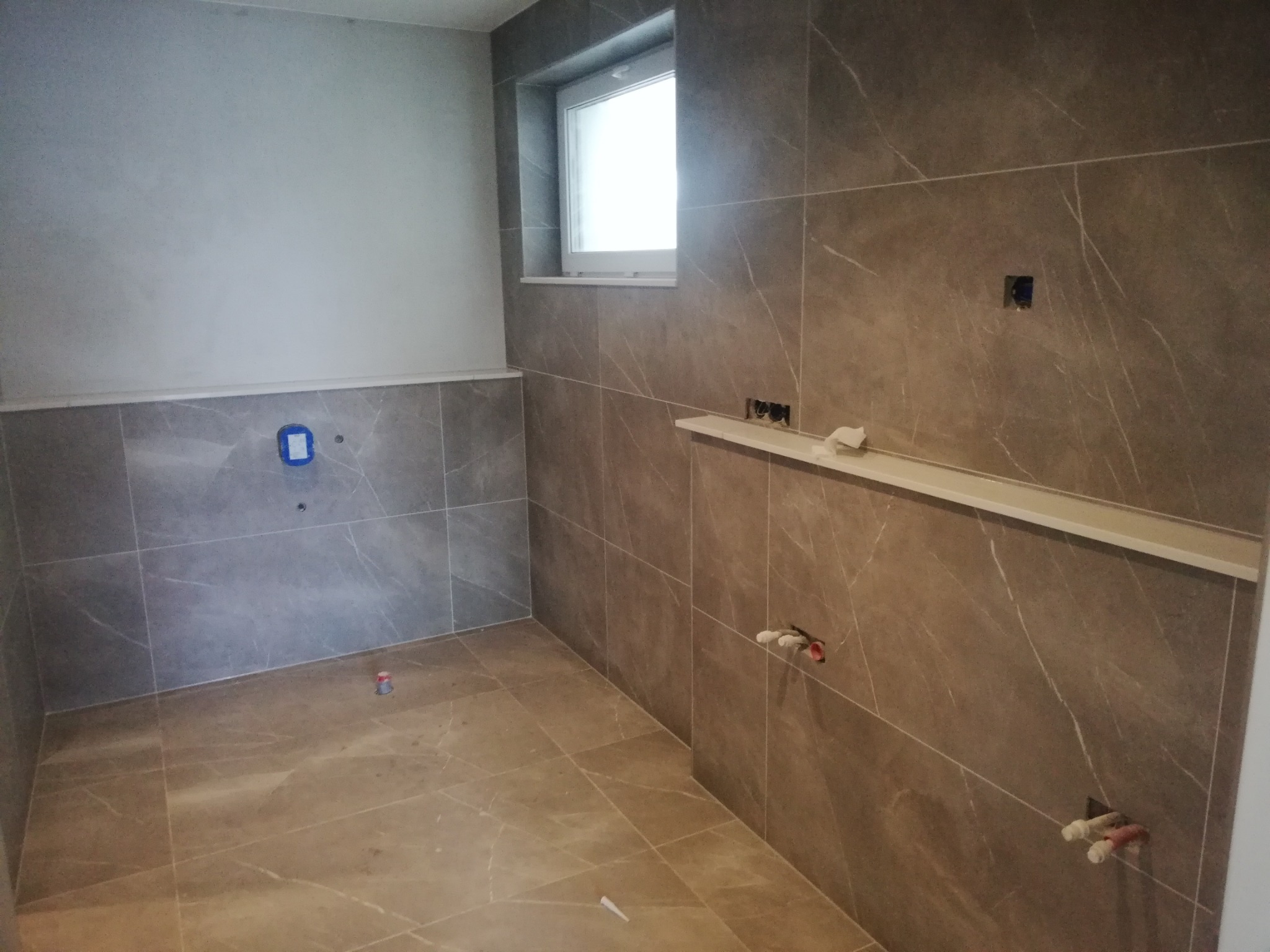 Szara łazienka w trakcie remontu, z marmurkowymi płytkami na ścianach i podłodze, widoczne przygotowane instalacje wodne i elektryczne oraz okno z parapetem.