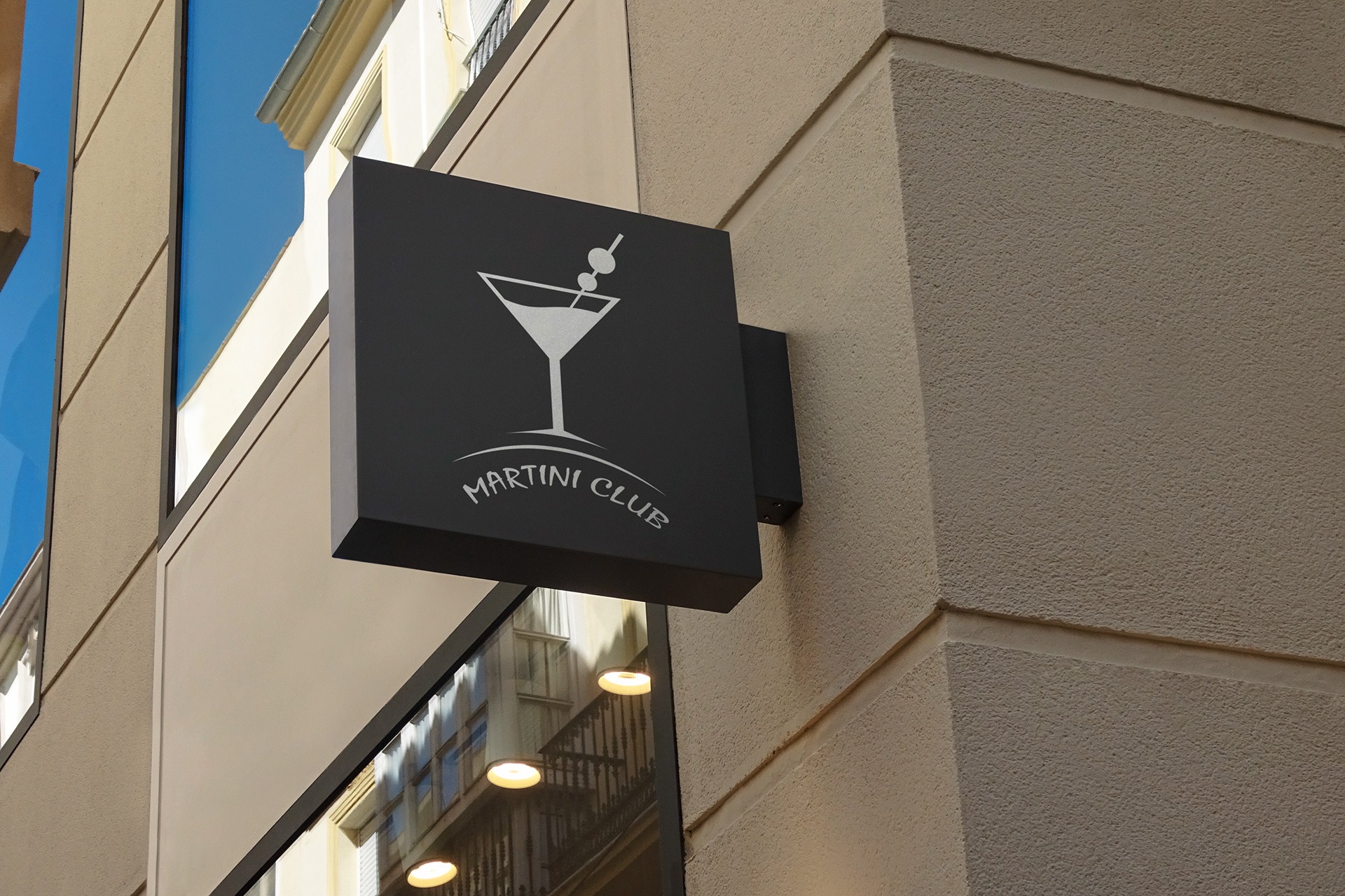 Czarny, kwadratowy szyld Martini Club z białym logo martini na elewacji budynku, widoczne odbicia w szybie okna.