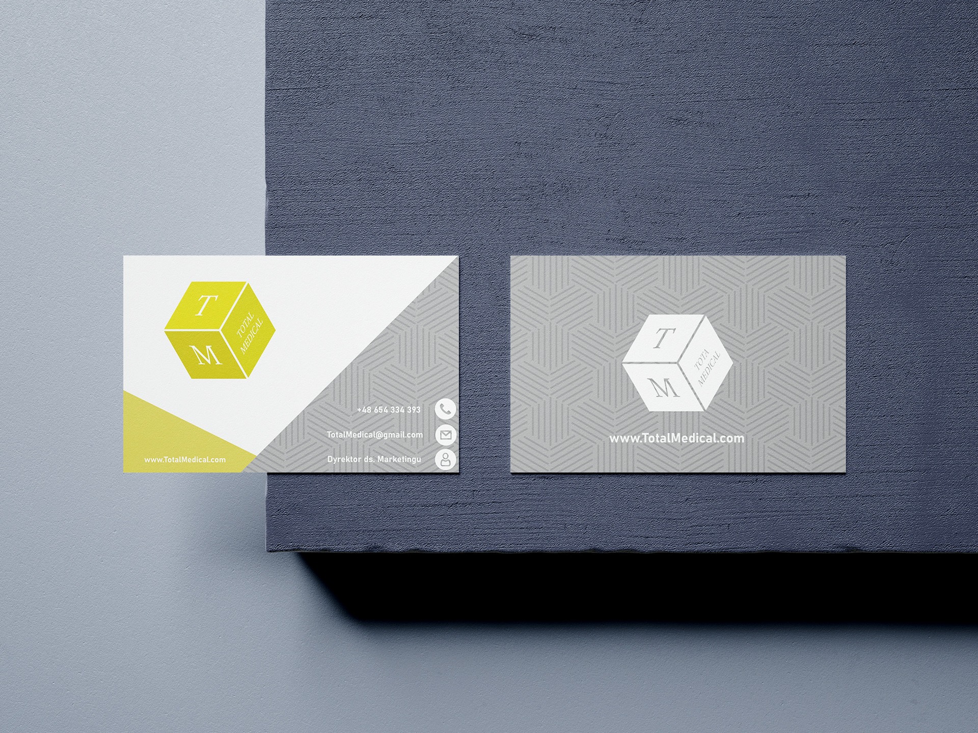 Dwie wizytówki firmy Total Medical na teksturowanym tle, jedna z żółto-białym designem, druga w odcieniach szarości, obie z logo w kształcie sześcianu z literami T i M.
