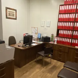 Wnętrze biura z biurkiem, komputerami, krzesłami i wysoką półką wypełnioną czerwonymi segregatorami, na ścianie wisi obrazek w ramce i dwa certyfikaty.