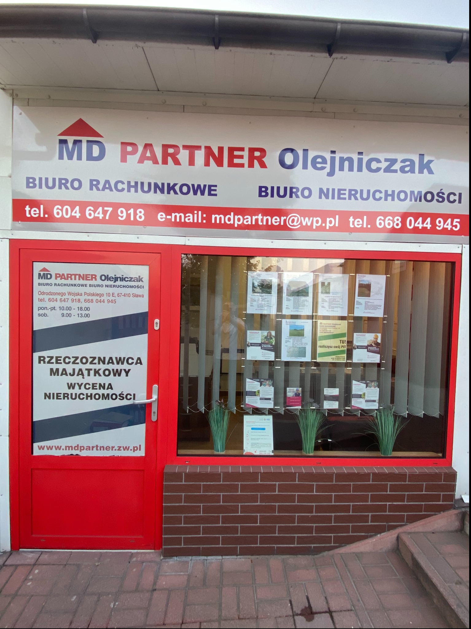 Fasada biura MD PARTNER Olejniczak w Sławie, z widocznymi informacjami o biurze rachunkowym i nieruchomości, adresie, numerach telefonów i adresie e-mail. Na drzwiach informacja o rzeczoznawcy...