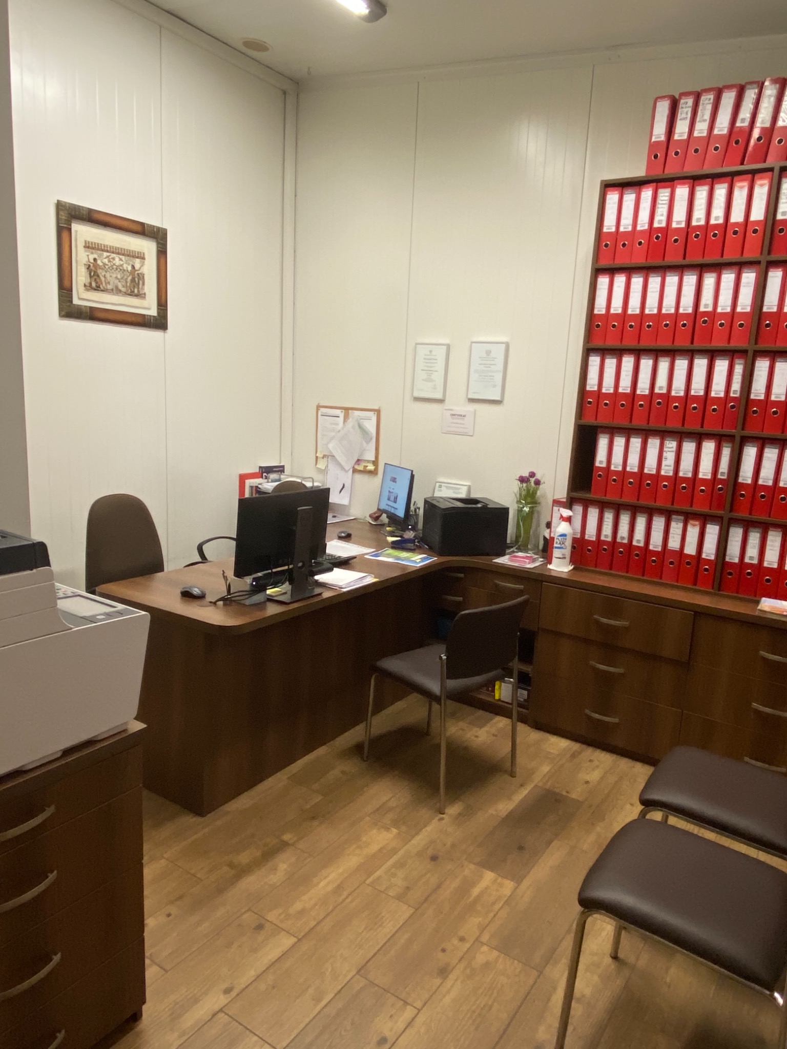 Wnętrze biura z biurkiem, komputerami, krzesłami i wysoką półką wypełnioną czerwonymi segregatorami, na ścianie wisi obrazek w ramce i dwa certyfikaty.