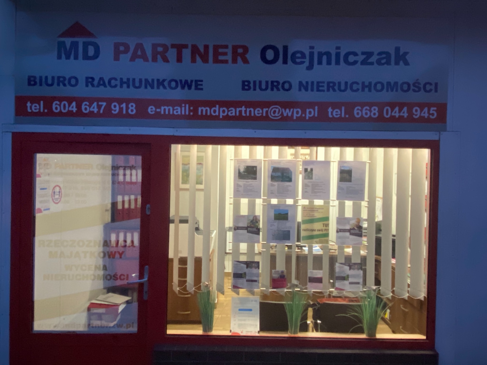 Fasada biura rachunkowego i nieruchomości MD Partner Olejniczak z oknami wystawowymi, na których wiszą ogłoszenia i ulotki informacyjne, widoczne z zewnątrz po zmroku.