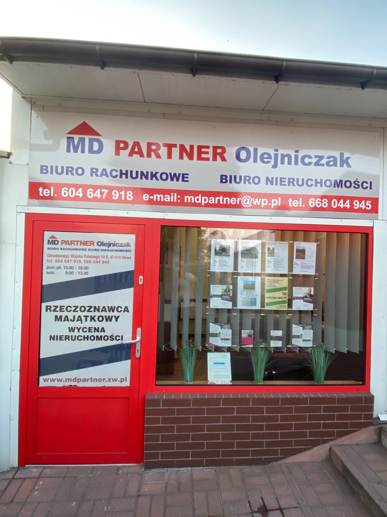 Fasada biura rachunkowego i nieruchomości MD Partner Olejniczak z czerwonymi drzwiami, witryną z ogłoszeniami i informacją o godzinach otwarcia, zlokalizowanego przy ulicy Odrodzonego Wojska...