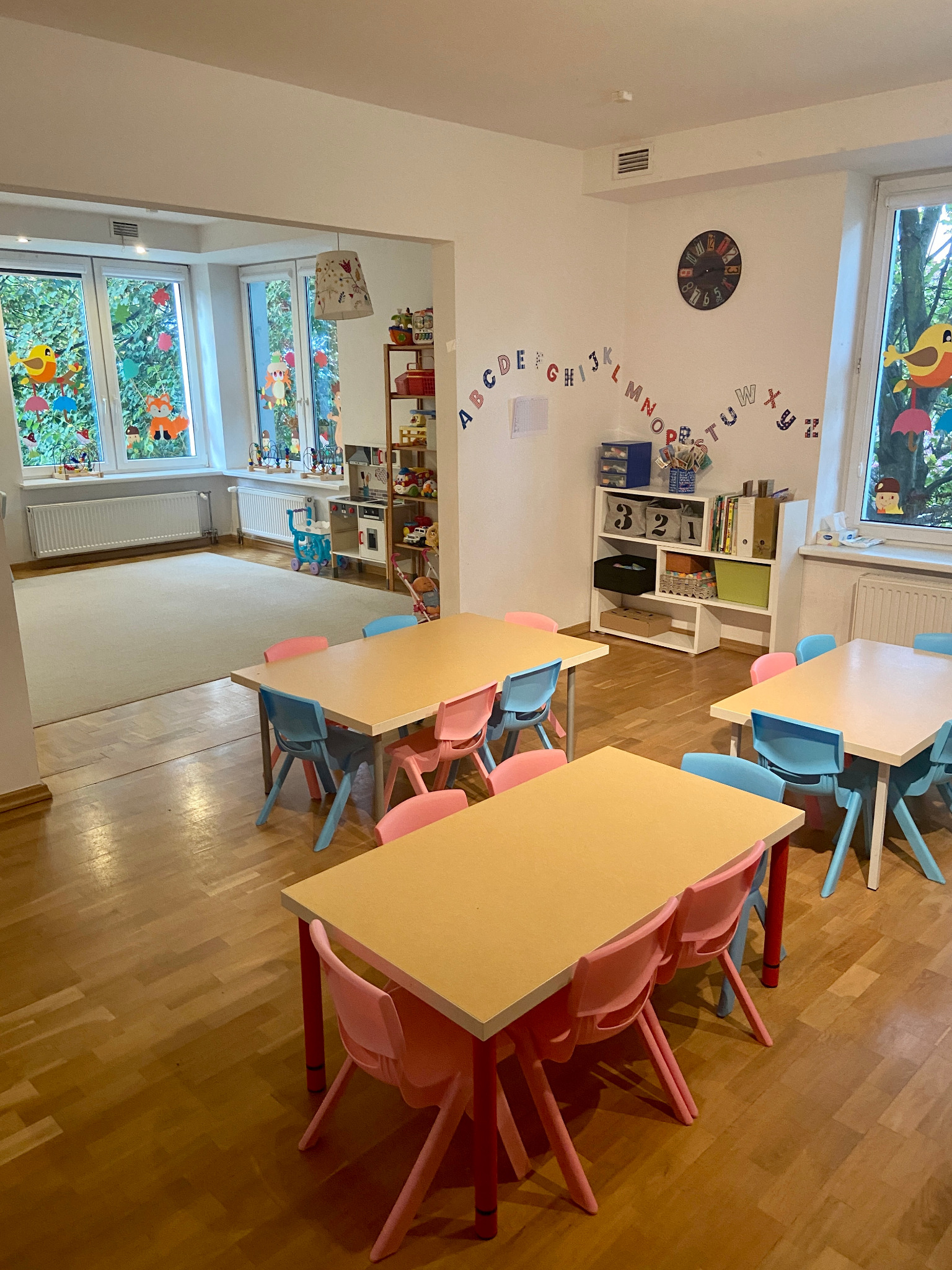Sala żłobkowa z drewnianą podłogą, małymi stolikami z kolorowymi krzesłami (różowe i niebieskie), dekoracjami okiennymi i alfabetycznymi na ścianie, oraz regałem z zabawkami w tle.