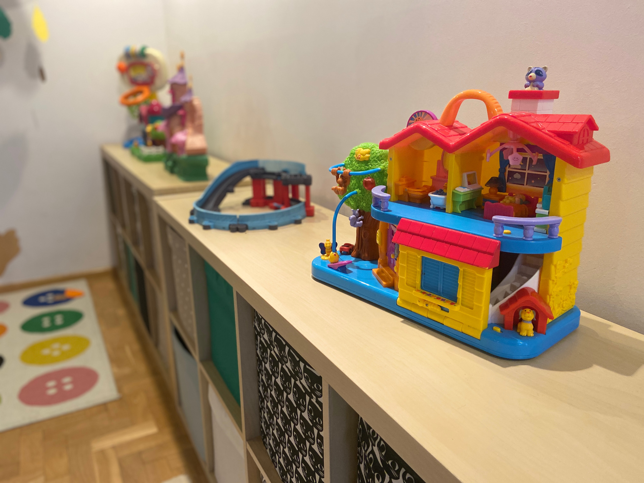 Kolorowy domek dla lalek z plastikowymi mebelkami i figurkami zwierząt na półce w pokoju dziecięcym, obok torów kolejowych i innych zabawek, dywan w guziki na podłodze.