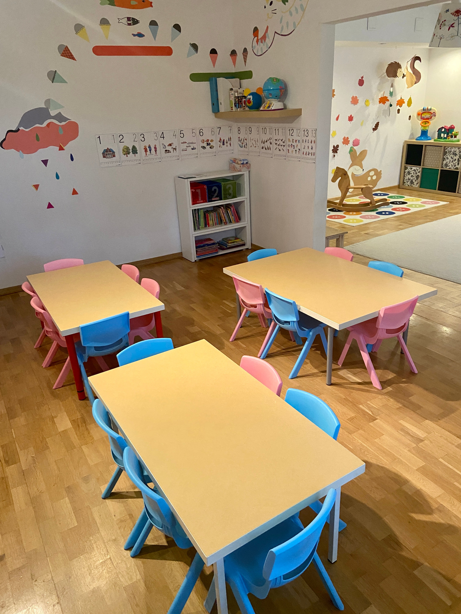 Sala zabaw dla dzieci z trzema drewnianymi stolikami otoczonymi niebieskimi i różowymi krzesełkami, dekoracje na ścianach w postaci naklejek z chmurką, lodami i cyframi, w tle regał z książkami...