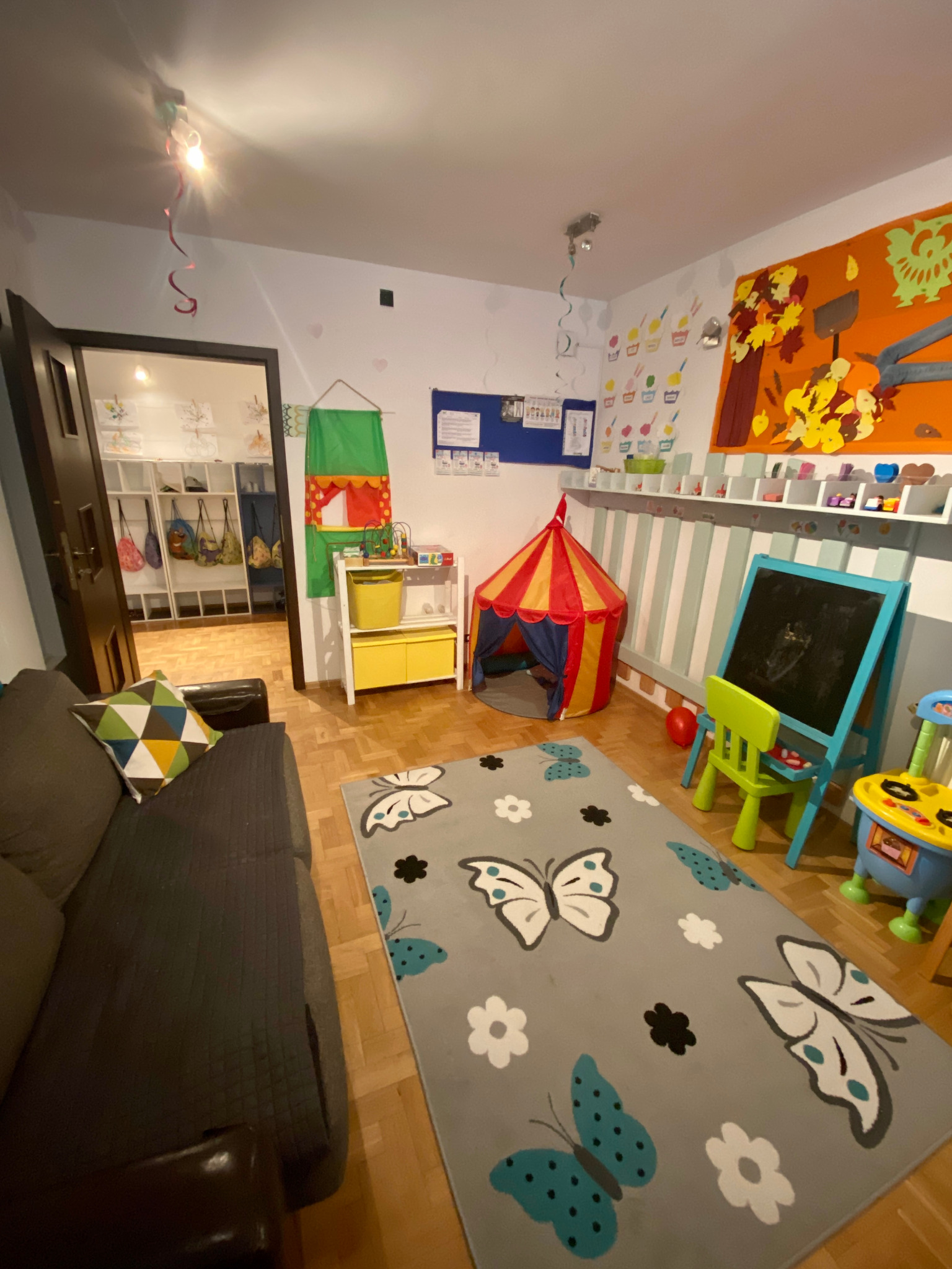 Pokój zabaw dla dzieci z szarą sofą z geometryczną poduszką, dywanem w motyle, namiotem tipi, tablicą, stolikiem edukacyjnym i dekoracjami na ścianach.