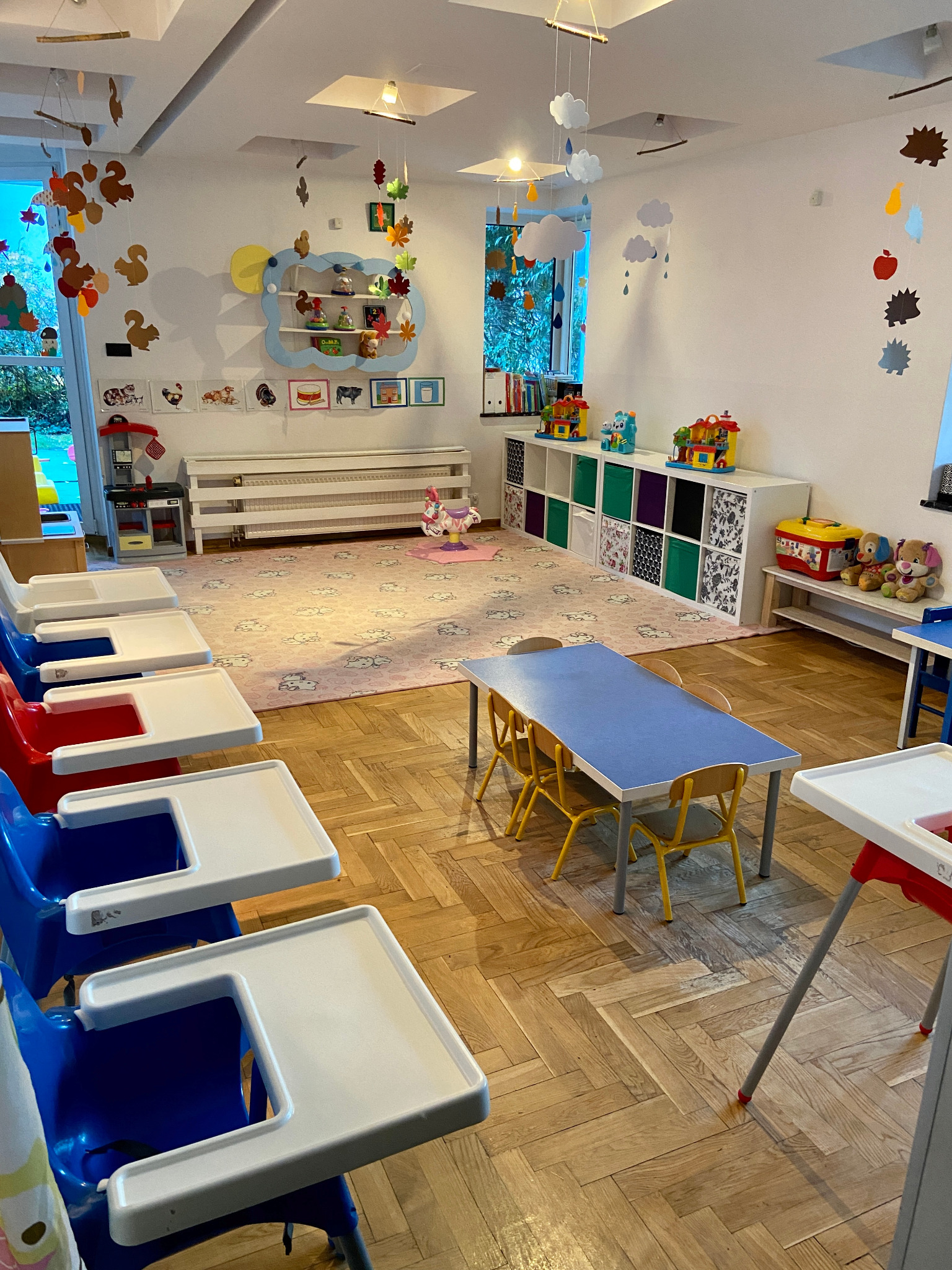 Jasne wnętrze sali zabaw z niskimi stolikami, krzesełkami i wysokimi krzesełkami do karmienia, dekoracje tematyczne jesienne i chmurkowe podwieszone pod sufitem, zabawki na półkach.