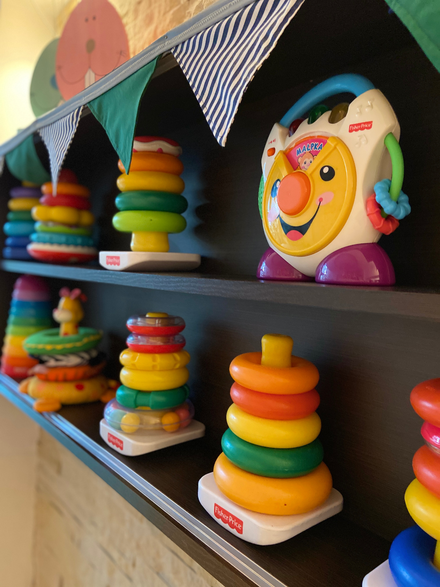 Półka z zabawkami dla dzieci: piramidki z kolorowych kółek Fisher-Price i interaktywna zabawka grająca z uśmiechniętą buzią, udekorowana girlandą z trójkątnych proporczyków.