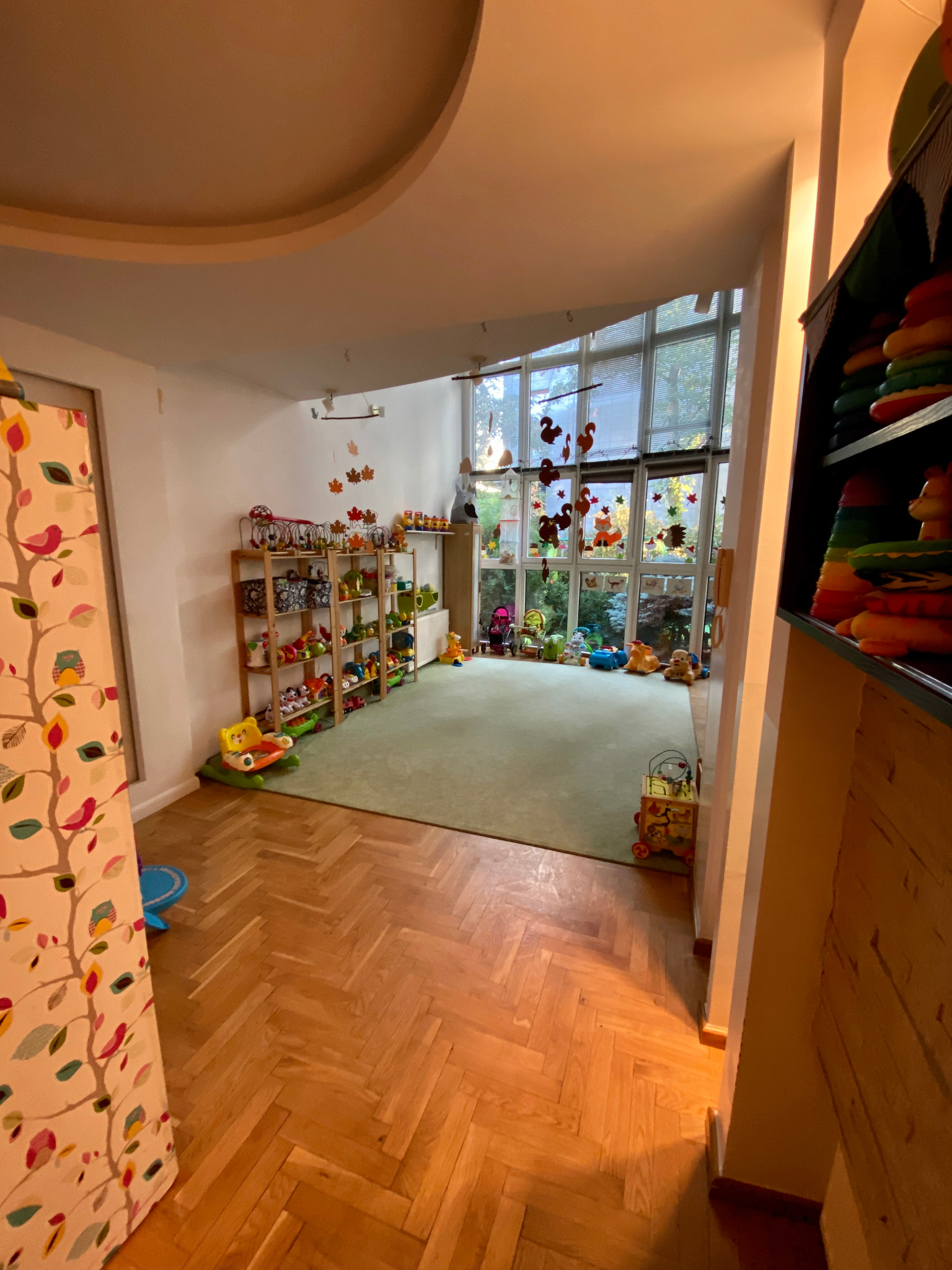 Sala zabaw w żłobku z drewnianą podłogą w jodełkę, zielonym dywanem i regałami pełnymi zabawek, okna zdobione jesiennymi dekoracjami.