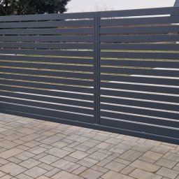 Usługi spawalniczo monterskie - Nowoczesna, przesuwna brama w kolorze grafitowym z poziomymi panelami, zamontowana na posesji z kostką brukową. Widoczny automat do bramy.