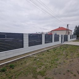 Usługi spawalniczo monterskie - Nowoczesne ogrodzenie posesji z poziomych, grafitowych paneli na jasnych, betonowych słupkach i podmurówce. Widok na posesję i dom w tle.