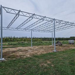 Usługi spawalniczo monterskie - Konstrukcja stalowa hali w budowie, widok z zewnątrz na szkielet z profili, tło stanowią drzewa i pochmurne niebo. Widoczne fundamenty pod słupy.
