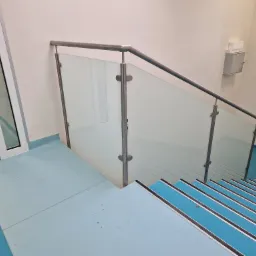 Wnętrze budynku: Schody z niebieskimi stopniami zabezpieczone balustradą ze szkła i metalu, widok z góry.