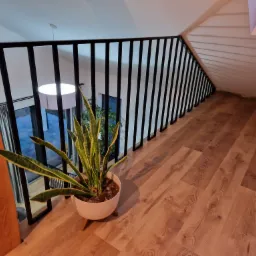 Nowoczesna balustrada metalowa w kolorze czarnym, z pionowymi szczeblami, zamontowana na drewnianej podłodze antresoli, z widokiem na okno i rośliną doniczkową w białej osłonce.