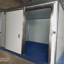 Usługi spawalniczo monterskie - Przesuwna brama przemysłowa z białych paneli, częściowo otwarta, widoczne metalowe prowadnice i niebieska podłoga wewnątrz, surowy mur z cegieł w tle.