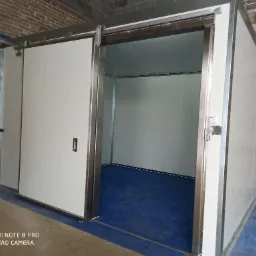 Przesuwna brama przemysłowa z białych paneli, częściowo otwarta, widoczne metalowe prowadnice i niebieska podłoga wewnątrz, surowy mur z cegieł w tle.