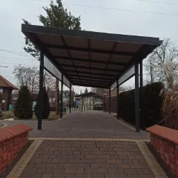 Zadaszenie wjazdu wykonane z konstrukcji stalowej, widok z perspektywy wjazdu na posesję, kostka brukowa w odcieniach brązu.