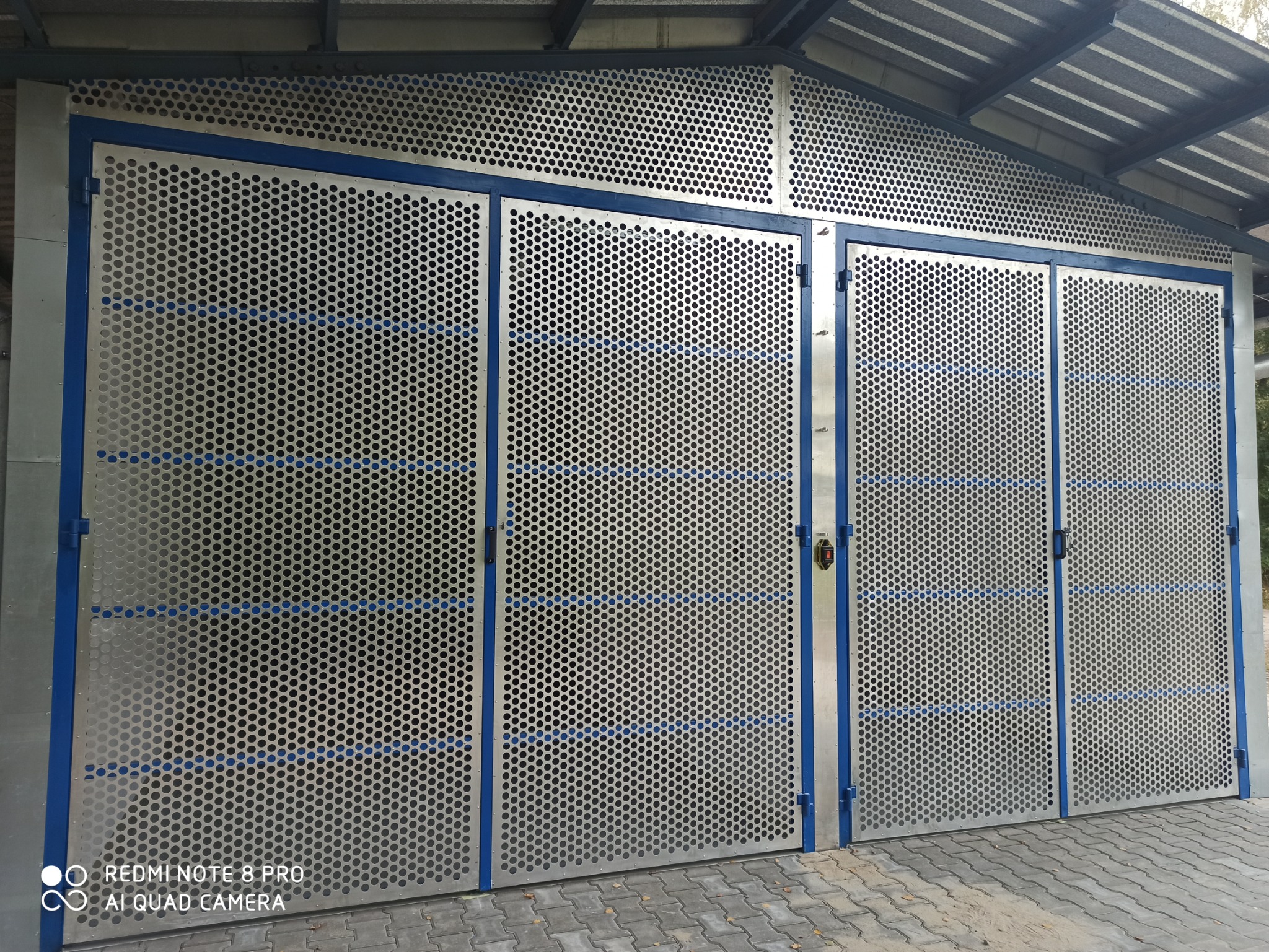 Metalowa konstrukcja z perforowanych paneli, rama w kolorze niebieskim, prawdopodobnie brama lub wrota do garażu/magazynu, widoczne zawiasy i zamki.