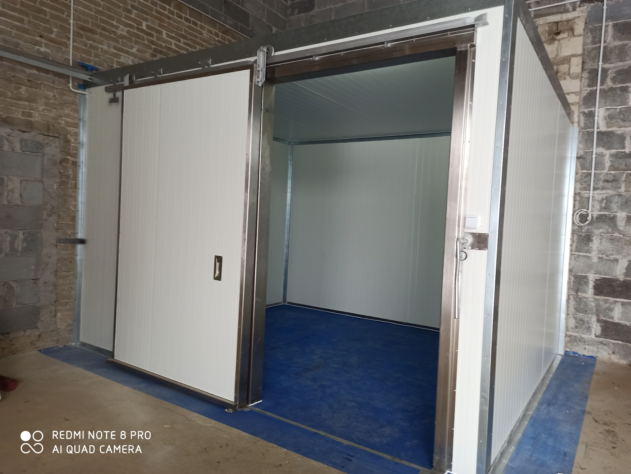 Przesuwna brama przemysłowa z białych paneli, częściowo otwarta, widoczne metalowe prowadnice i niebieska podłoga wewnątrz, surowy mur z cegieł w tle.