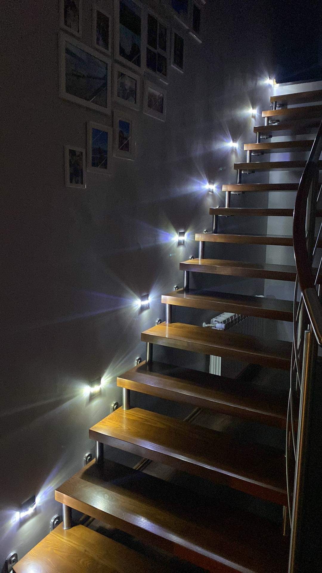 Drewniane schody z podświetleniem LED na ścianie, zdjęcia w ramkach na ścianie, metalowa barierka.
