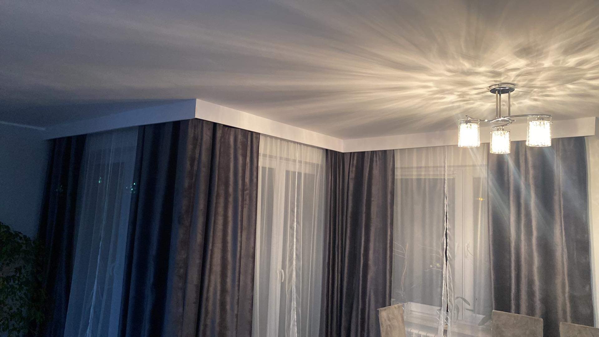 Zasłony welurowe grafitowe z białymi firanami, karnisz sufitowy z listwą maskującą, kryształowy żyrandol rzucający wzory na sufit.