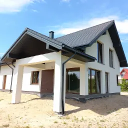 Nowoczesny dom jednorodzinny z białymi ścianami i grafitowym dachem, widok z zewnątrz, teren wokół domu w trakcie prac ziemnych.