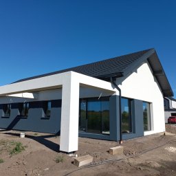 Szymon Tadzik - Nowoczesny dom jednorodzinny z płaskim dachem, białą elewacją i szarymi akcentami, widok z poziomu gruntu na tle błękitnego nieba, teren wokół domu w trakcie prac wykończeniowych.