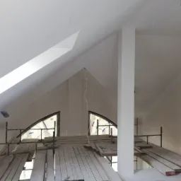 Poddasze w trakcie remontu z widocznymi rusztowaniami, deskami roboczymi, oknami w kształcie łuków zabezpieczonymi folią i świeżo pomalowanymi ścianami na biało.