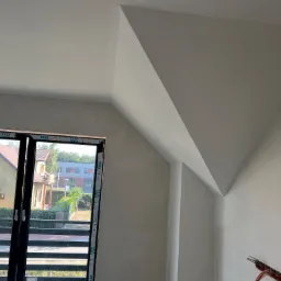 Poddasze po gładziowaniu, widok przez okno na dom. Ściany i sufit w jasnym odcieniu, widoczne niedoskonałości przed malowaniem. Okno z czarną ramą.