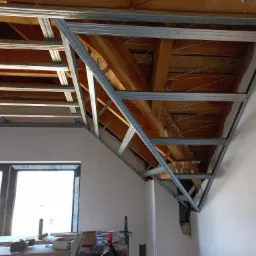 Skelet konstrukcji sufitu podwieszanego z profili metalowych, widoczne drewniane belki stropowe, okno w tle. Prace wykończeniowe w nowym budynku.