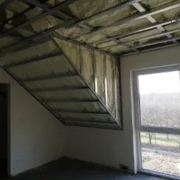 Poddasze w trakcie ocieplania, widoczna konstrukcja metalowa i wełna mineralna, okno z widokiem na drzewa bez liści, surowy stan wykończenia.