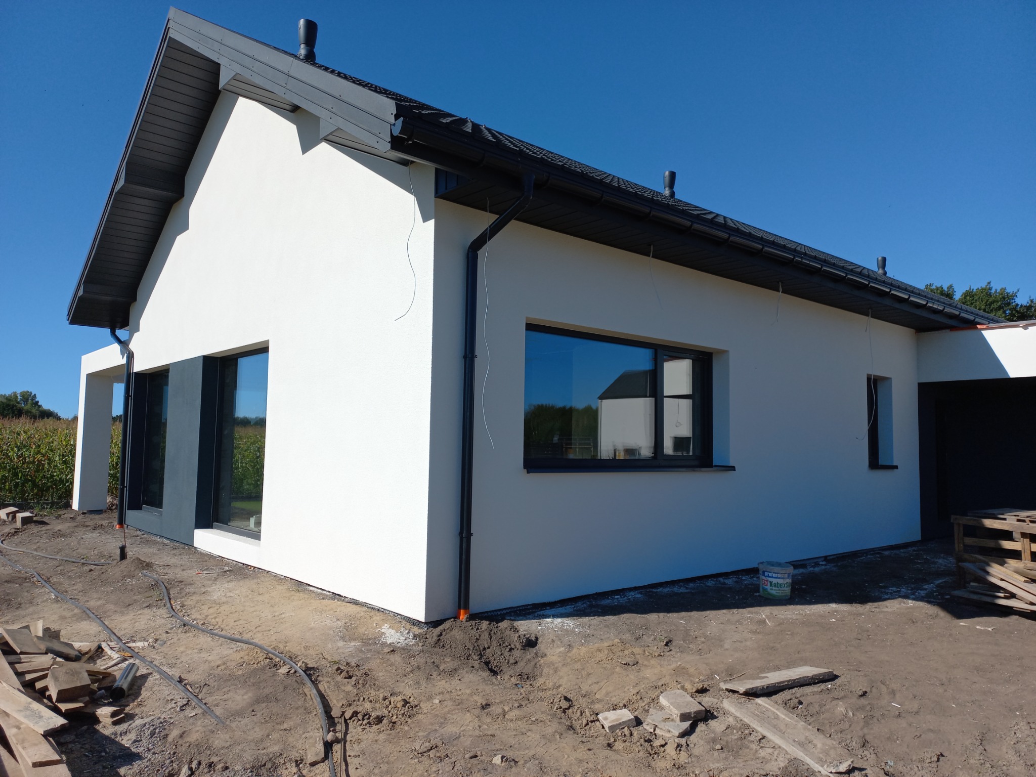 Nowoczesny dom jednorodzinny z białą elewacją, czarną stolarką okienną i dachem, widoczne elementy wykończeniowe i teren wokół budynku w trakcie prac.