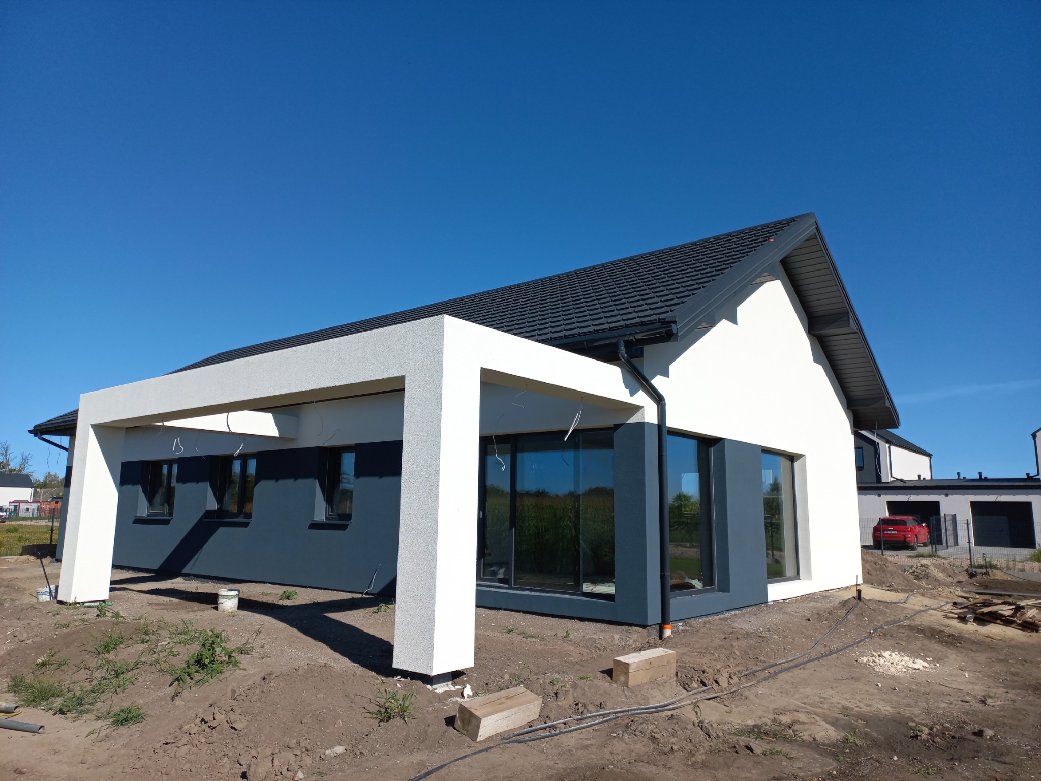 Nowoczesny dom jednorodzinny z płaskim dachem, białą elewacją i szarymi akcentami, widok z poziomu gruntu na tle błękitnego nieba, teren wokół domu w trakcie prac wykończeniowych.