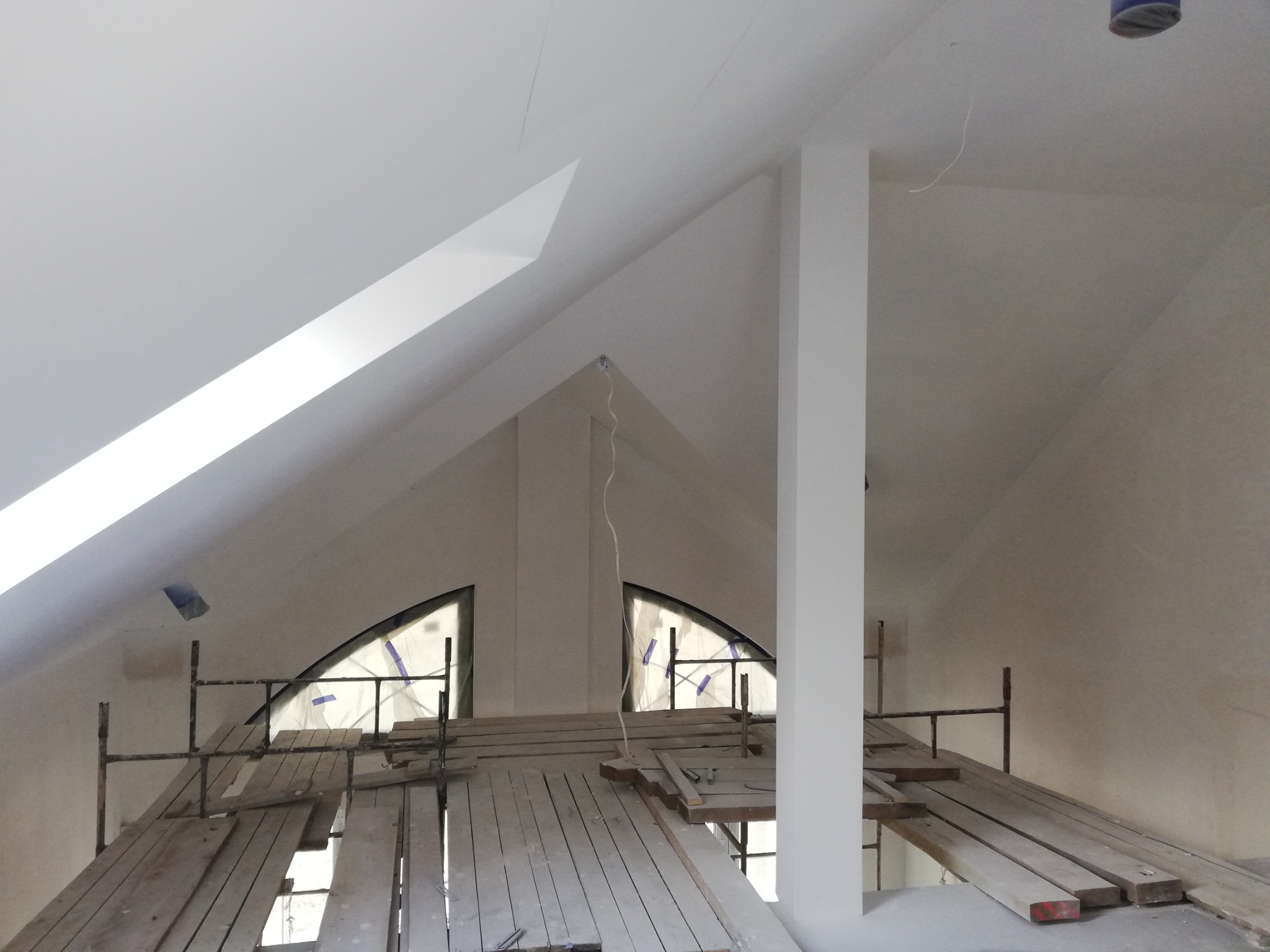 Poddasze w trakcie remontu z widocznymi rusztowaniami, deskami roboczymi, oknami w kształcie łuków zabezpieczonymi folią i świeżo pomalowanymi ścianami na biało.