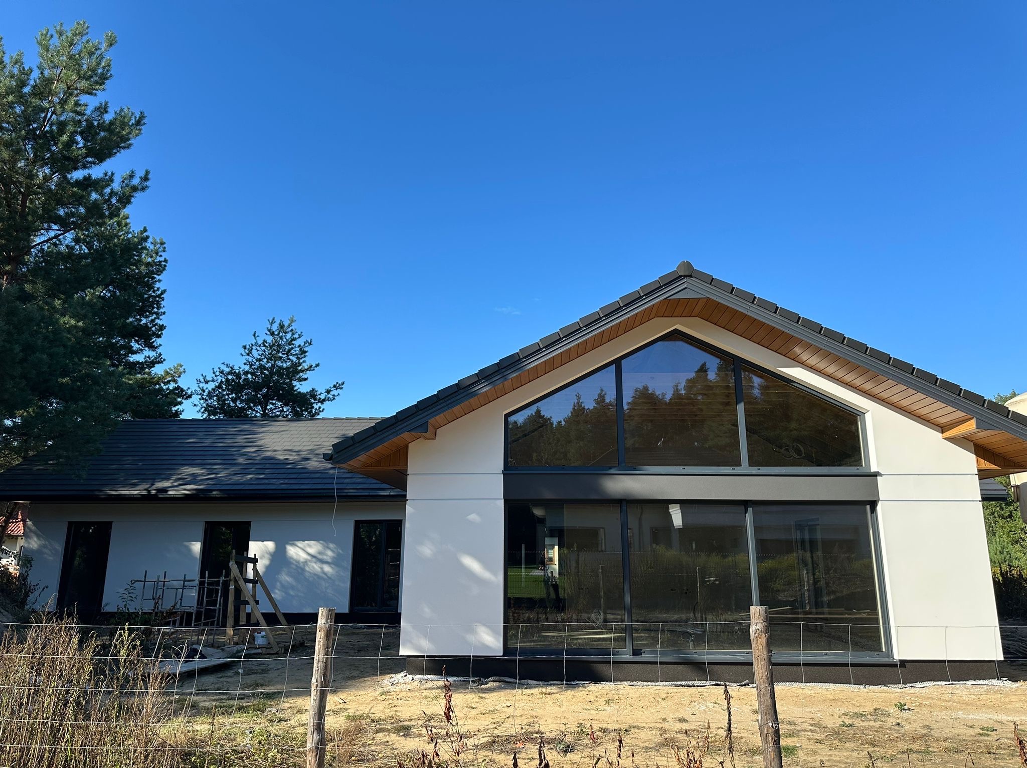 Nowoczesny dom z białą elewacją i dużymi oknami, widoczny fragment rusztowania. Dom otoczony drzewami, w tle błękitne niebo.