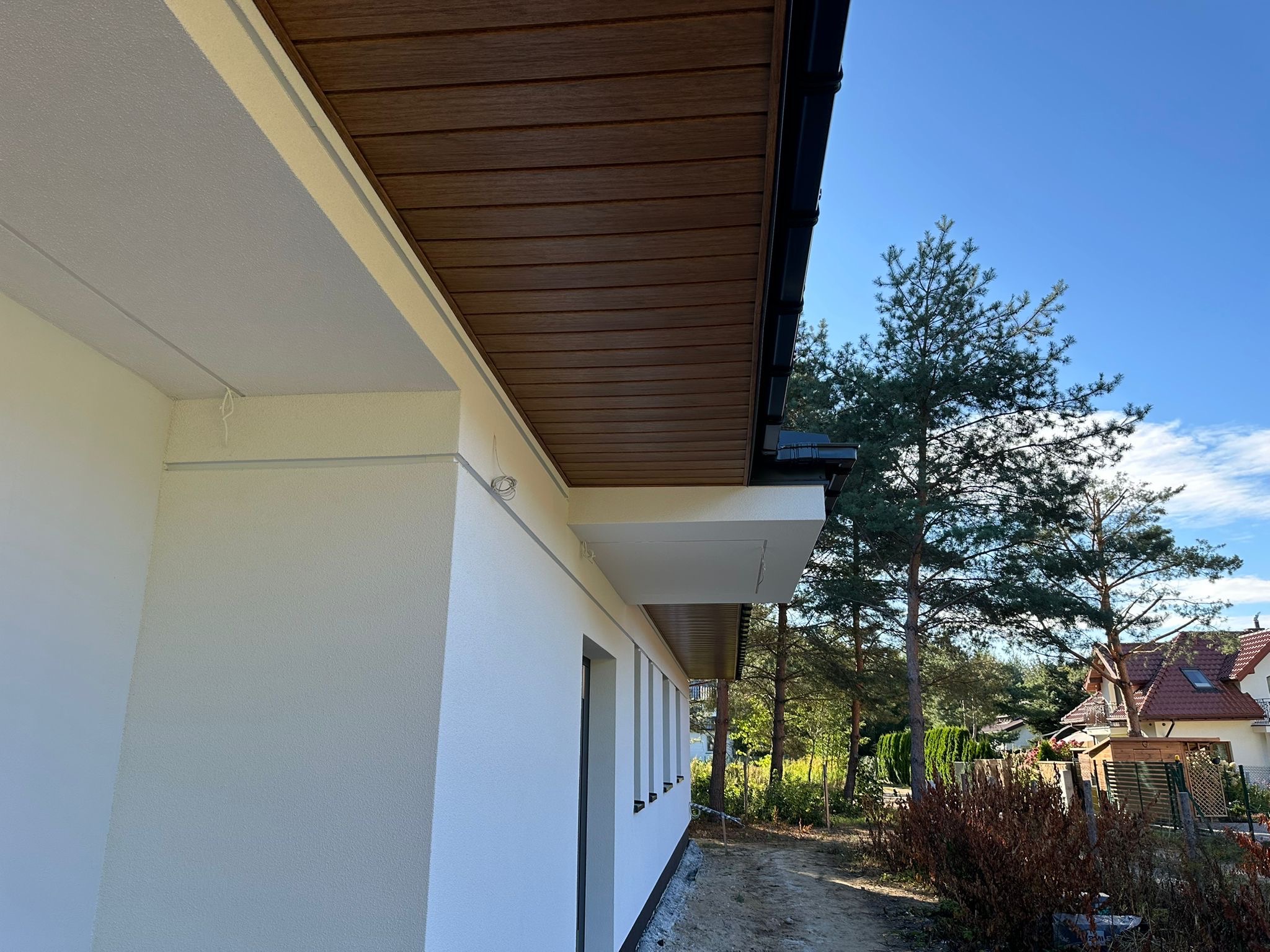 Jasna elewacja domu jednorodzinnego z drewnopodobną podbitką dachową, widok z boku, w tle drzewa i fragment sąsiedniego budynku.