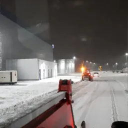 Widok z kabiny odśnieżarki na odśnieżaną drogę w nocy, w tle budynki przemysłowe i światła uliczne.