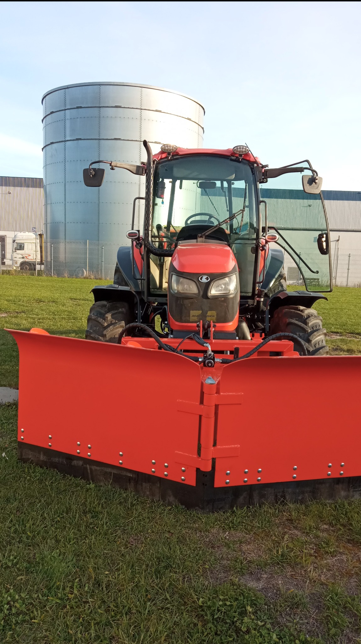 Czerwony traktor z pługiem do odśnieżania stoi na trawie, w tle silos i budynek przemysłowy.