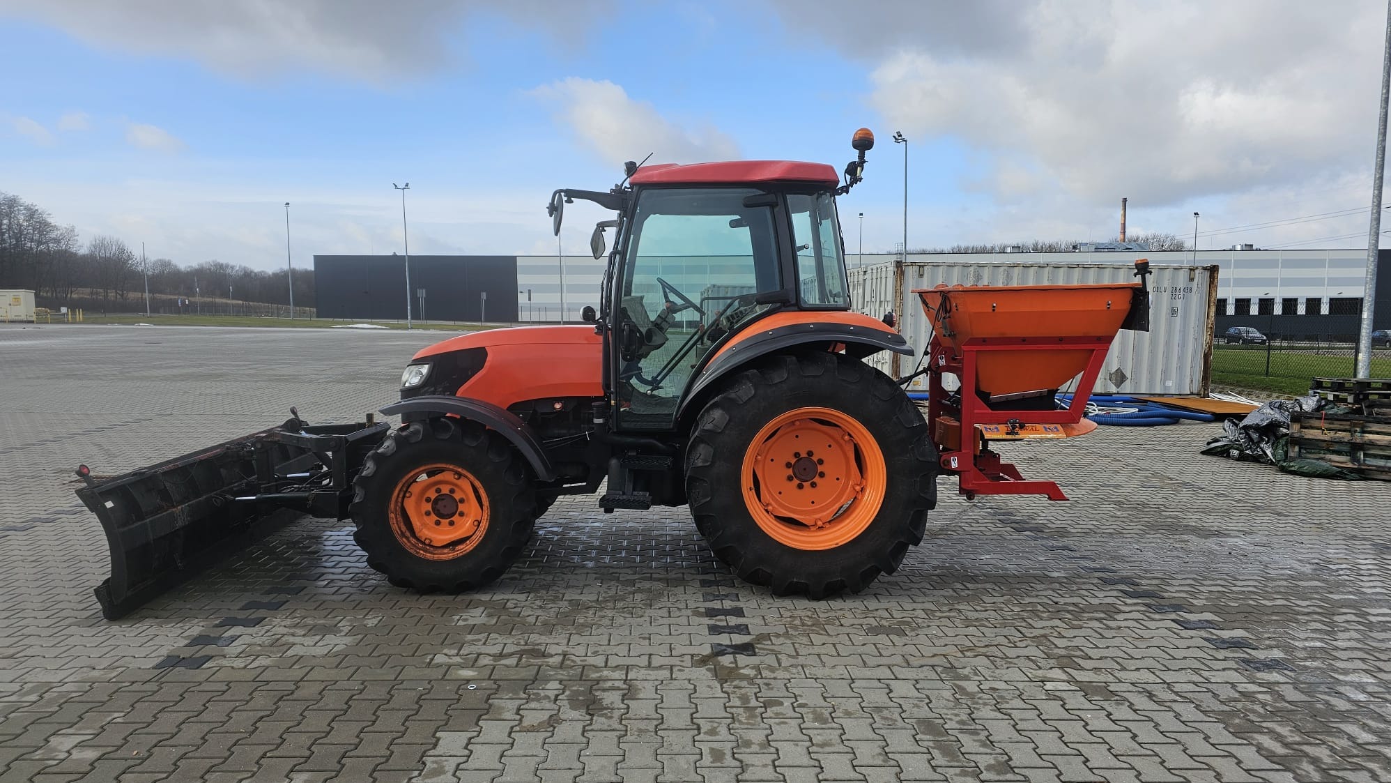 Pomarańczowy traktor z pługiem i rozsiewaczem soli na kostce brukowej, w tle budynki magazynowe i pochmurne niebo.