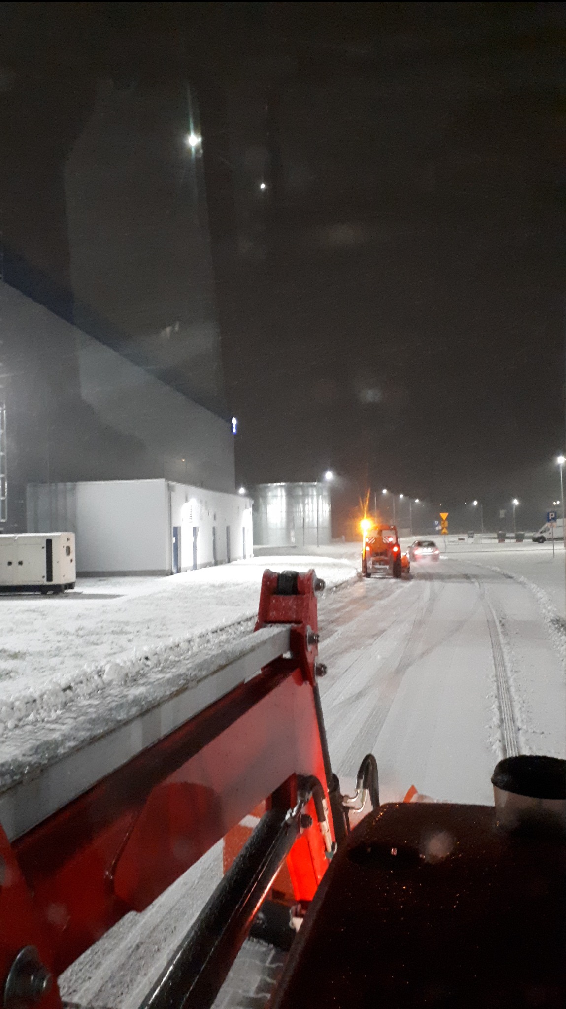 Widok z kabiny odśnieżarki na odśnieżaną drogę w nocy, w tle budynki przemysłowe i światła uliczne.