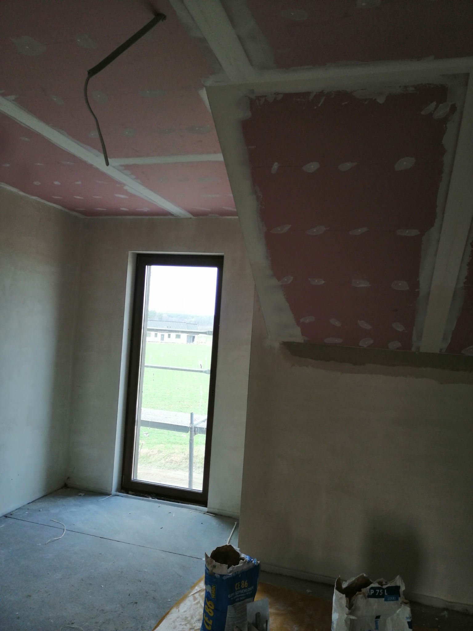 Poddasze w trakcie wykończenia: widoczne płyty kartonowo-gipsowe na skośnym suficie, ściany po wstępnym tynkowaniu, okno z widokiem na zewnątrz oraz worki z zaprawą na podłodze.