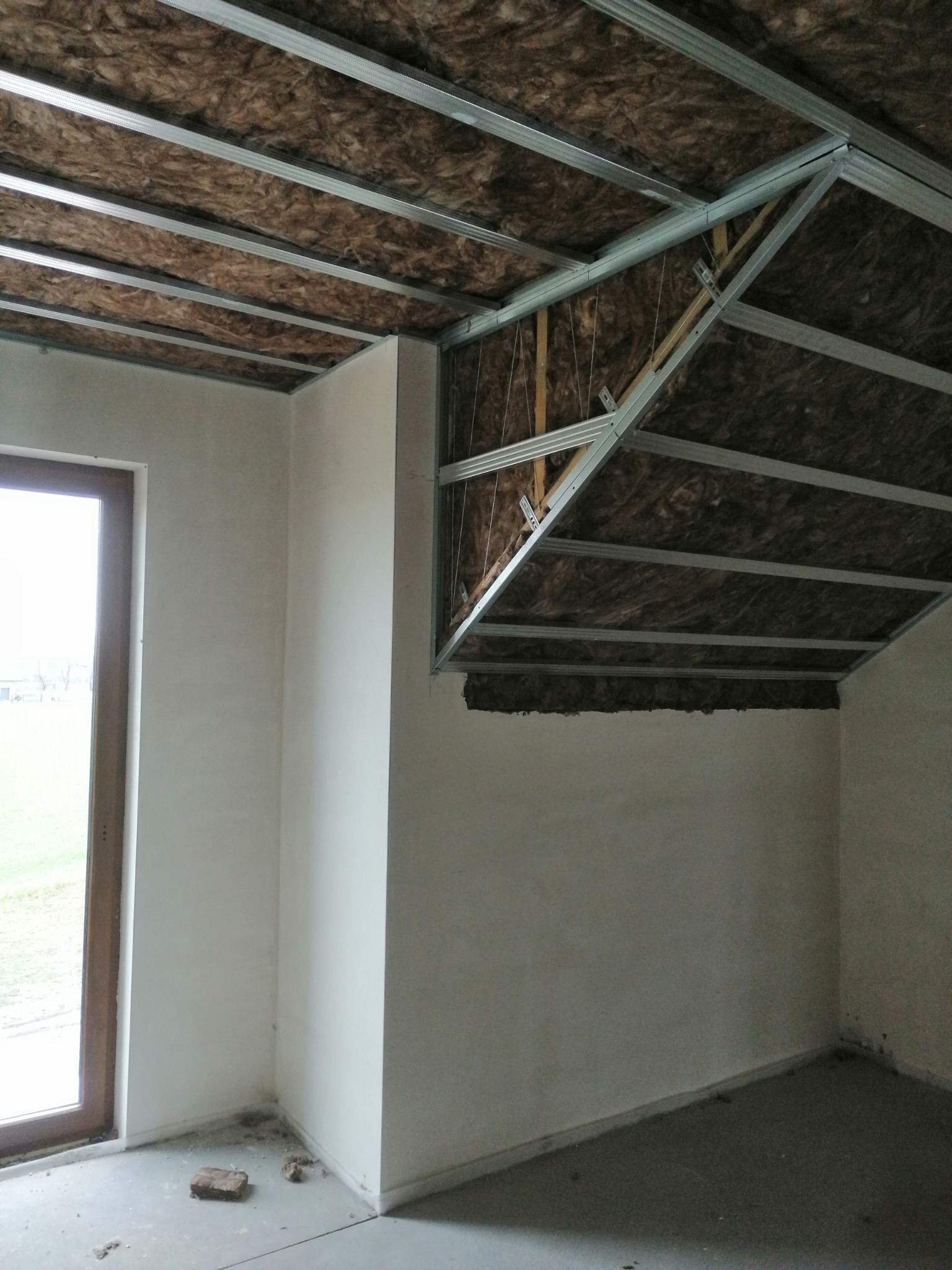 Poddasze w trakcie ocieplania, widoczna wełna mineralna między metalowymi profilami konstrukcyjnymi, surowe ściany i okno z drewnianą ramą.