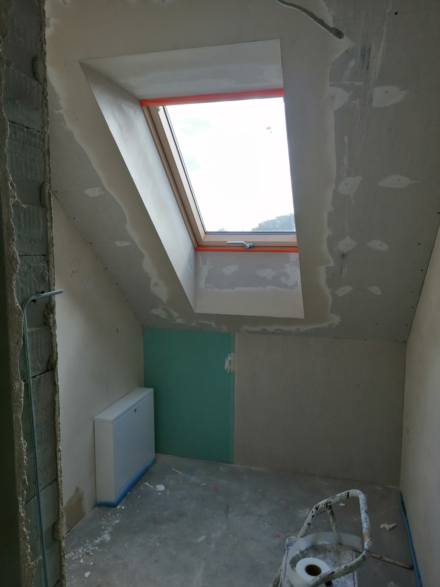 Poddasze w trakcie ocieplania z zamontowanym oknem dachowym, widoczne płyty gipsowo-kartonowe, zabezpieczone okno i drabina malarska.