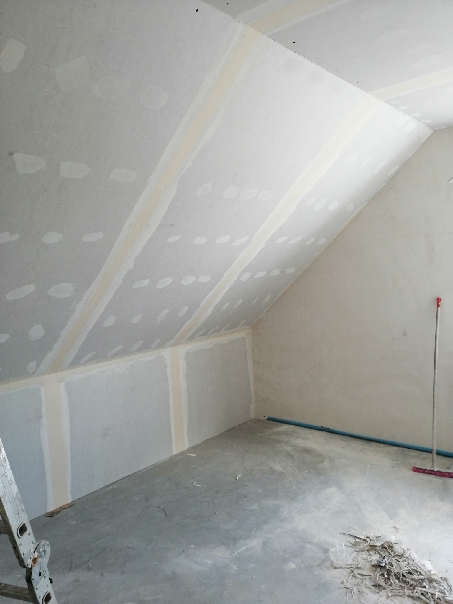 Poddasze w trakcie wykańczania: widoczne płyty gipsowo-kartonowe na skosach i ścianach, z widocznymi łączeniami wymagającymi szpachlowania, fragment drabiny po lewej stronie i niebieska rura przy...