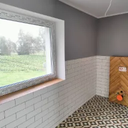 Wnętrze łazienki w trakcie remontu z białą cegiełką na ścianach do połowy wysokości, szarą górną częścią ściany, wzorzystą podłogą, oknem z widokiem na zieleń i obudową stelaża WC z drewnianych...