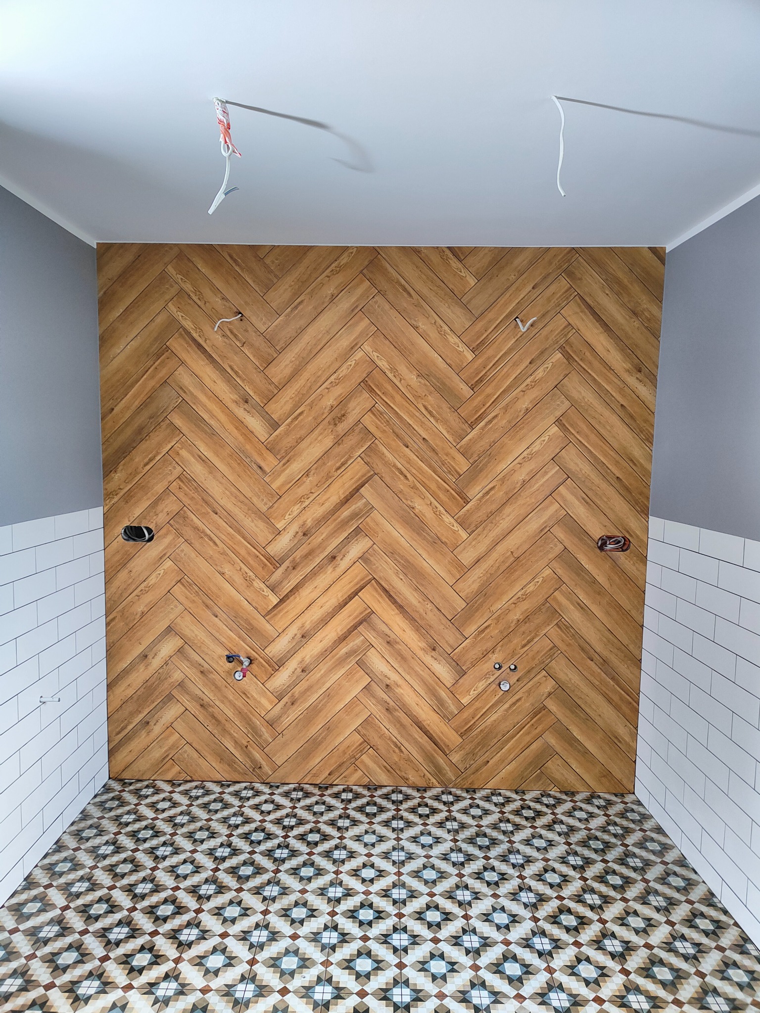 Łazienka w trakcie remontu: drewniana ściana ułożona w jodełkę, białe płytki na ścianach do połowy wysokości, wzorzysta podłoga, wystające kable i rury.