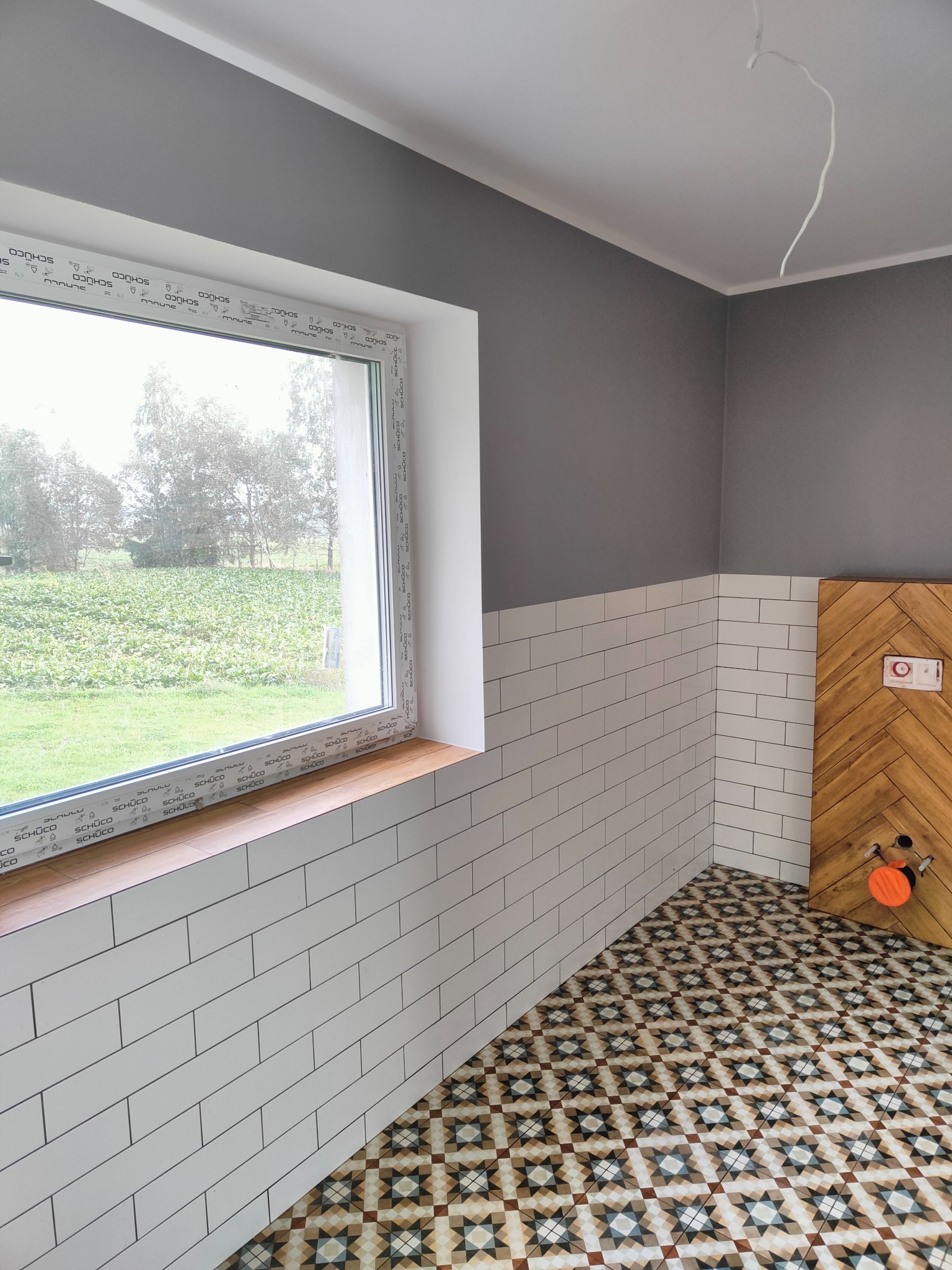 Wnętrze łazienki w trakcie remontu z białą cegiełką na ścianach do połowy wysokości, szarą górną częścią ściany, wzorzystą podłogą, oknem z widokiem na zieleń i obudową stelaża WC z drewnianych...